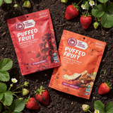 Two packages of puffed fruit snacks on a soil background with strawberries around.